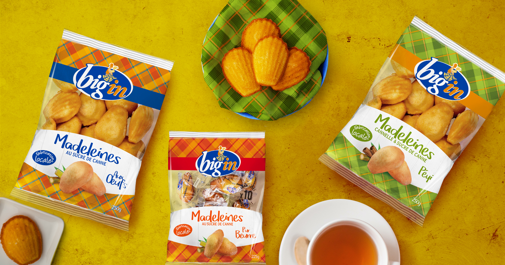 Photo instant de consommation sachets de madeleines Big’In, vue de dessus, sur un plan de travail jaune avec une tasse de thé, un bol avec des madeleines